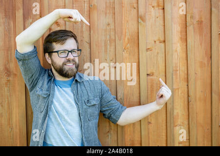 Cute, smiling bearded Caucasian man in glasses shows a gesture with index fingers on copy space, on a painted, old wooden wall. Close-up. Stock Photo