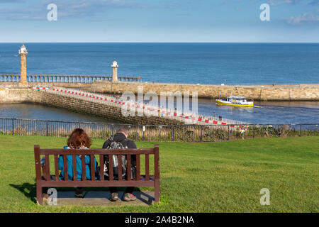 A pleasure cruiser leaves Whitby Harbour viewed by a couple seated on the high promenade of West Cliff Stock Photo