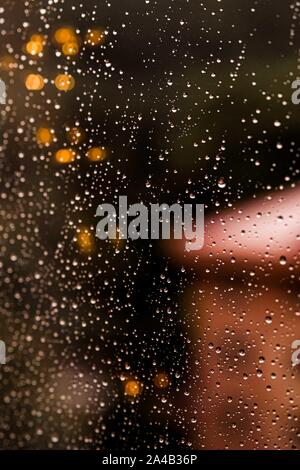 Drops Of Rain On the Glass with autumn city background. Warm Bokeh defocused Lights inside the house.. Abstract Backdrop. Vertical format. Stock Photo