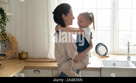Happy mother holding little daughter, hugging, standing in kitchen Stock Photo