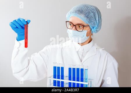 A female scientist, a pharmacist, a medic with glasses, is conducting an experiment in a laboratory, holding a test tube with red liquid in his hands.