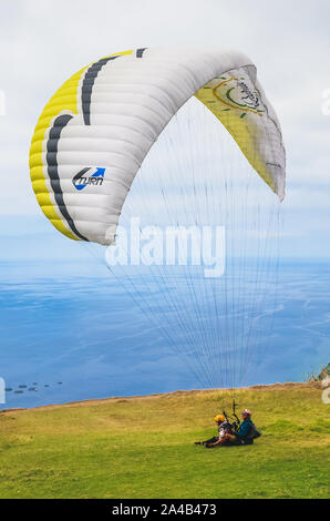 Arco da Calheta, Madeira, Portugal - Sep 16, 2019: Tandem paragliders landing on the cliffs above the Atlantic ocean. The blue seawater in the background. Paragliding, extreme sports. Stock Photo