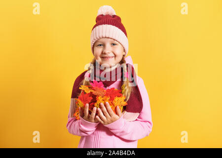 Cute cheerful little girl with an armful of autumn maple leaves. Child in hat and scarf on a yellow background. Autumn concept. Stock Photo