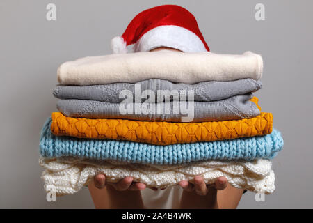 Woman in santa hat holding a stack of warm knitted sweaters on a gray wall background. Winter and christmas concept. Stock Photo