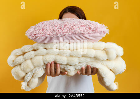 Woman holding a stack of warm fluffy plaid and pillows on a background of yellow wall. The concept of winter and autumn. Stock Photo