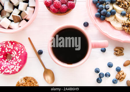 Tasty pink pastry, healthy morning breakfast, granola with banana and coconut pieces. A cup of fresh coffee on white wooden background. Stock Photo