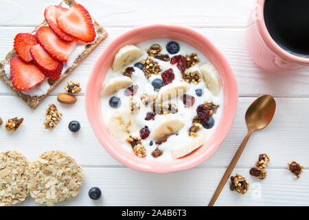 A bowl of fresh yougurt with granola, banana abd blueberry and a tasty fruit sandwich on white wooden table, top view. Stock Photo