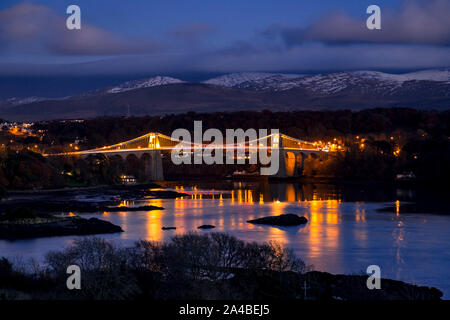 Thomas Telford suspension bridge across the Menai Straits at night, with snow capped mountains of Snowdonia in the background Stock Photo