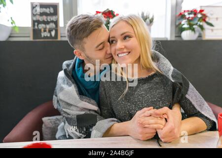 Kissing embracing young couple sitting in cafe together under one warm blanket Stock Photo
