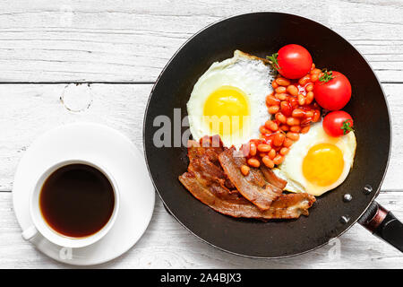 Full english breakfast in frying pan with fried eggs, bacon, beans, tomatoes and coffee on white wooden background. top view Stock Photo