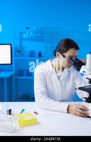 Portrait of young female scientist looking in microscope while doing research in medical laboratory lit by blue light, copy space Stock Photo