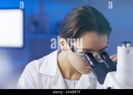 Closeup portrait of young female scientist looking in microscope while doing research in medical laboratory lit by blue light, copy space Stock Photo