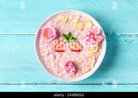 overnight oats with fresh strawberries, almonds and mint in a bowl with rose flowers on blue wooden table. healthy breakfast. top view Stock Photo
