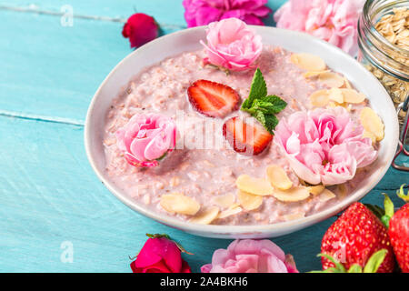 overnight oats with fresh strawberries, almonds and mint in a bowl with rose flowers on blue wooden table. healthy breakfast. close up Stock Photo