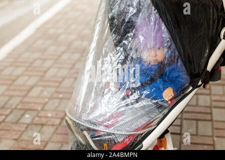 Little cute caucasian toddler boy in casual sport clothes having fun enjoy walking during rain in autumn day. Child sitting in stroller with rain Stock Photo