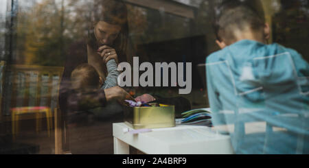 View through the window of a young mother breastfeeding her daughter next to her son drawing and creating. Stock Photo