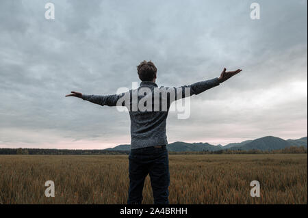 View from behind of a young man standing in autumn meadow under cloudy evening sky with his arms wide open. Stock Photo