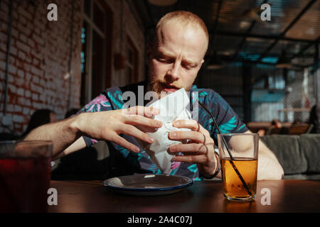 The guy holds a juicy hamburger in a summer cafe and lemonade on the table Stock Photo