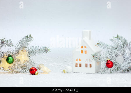 winter holidays ornaments against white snow background with christmas tree branches, decorative glass house and glowing star lights Stock Photo