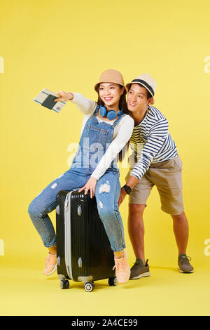 Young Vietnamese man pushing suitcase when his girlfriend sitting on it and pointing with hand holding passports and plane tickets Stock Photo