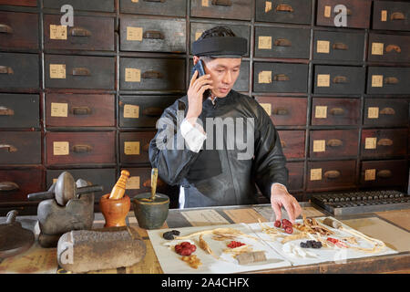 Traditional pharmacy worker talking on phone with customer when packing order of dry herbs, roots and berries Stock Photo
