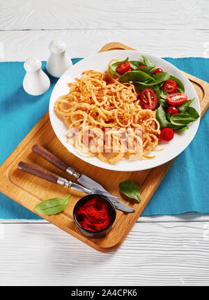 fresh spinach and tomato salad served with french crispy fried onion rings and strings on a white plate on a wooden board, vertical view from above Stock Photo