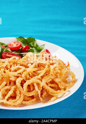 crispy fried onions with fresh spinach and tomato salad on a white plate on a textile table mat, vertical view Stock Photo