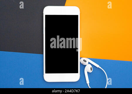 Music, gadgets, music lover. White smartphone and headphones on the black, yellow and blue backgrounds with headphones. View from above. Flat lay, top Stock Photo