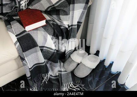 Woolen warm blanket and red book on white leather armchair, cozy home slippers on wooden floor. Relaxing weekend and stay at home concept. Copy space. Stock Photo