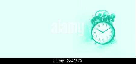 Hand holding black alarm clock through hole in mint color background. Wake up alert concept. Trendy green and turquoise color. Morning routine. Banner Stock Photo
