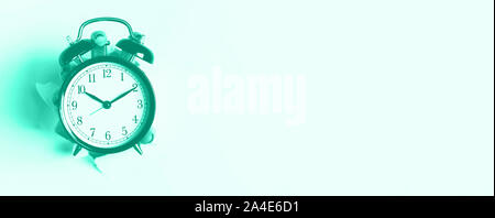 Hand holding black alarm clock through hole in mint color background. Wake up alert concept. Trendy green and turquoise color. Morning routine. Banner Stock Photo