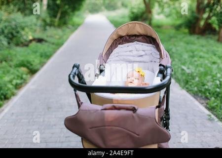 Cute little baby girl in yellow dress in brown baby carriage at outdoor, spring or summer season with fresh green background. Stock Photo