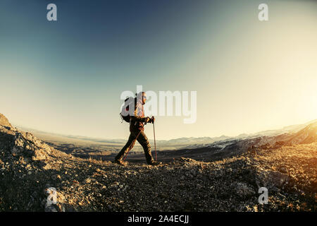 Hiker walks with backpack and trekking poles in sunset mountains Stock Photo