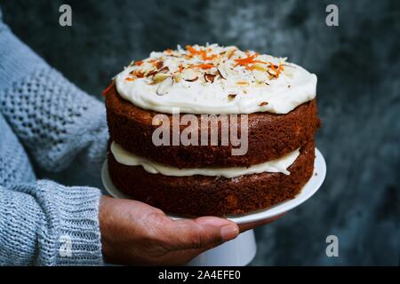 Hand holding cake stand with homemade carrot cake  cream cheese frosting on dark moody setting, selective focus Stock Photo