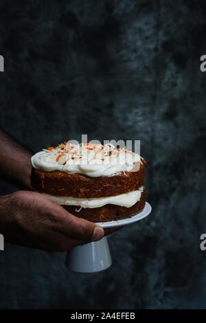 Hand holding cake stand with homemade carrot cake  cream cheese frosting on dark moody setting, selective focus Stock Photo