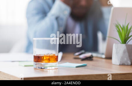 Desperate black businessman suffering from bankruptcy, drinking alcohol at workplace. Stock Photo