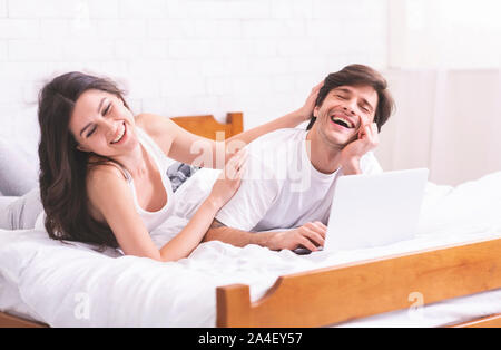 Young millennial couple laughing, getting crazy in bed with laptop Stock Photo