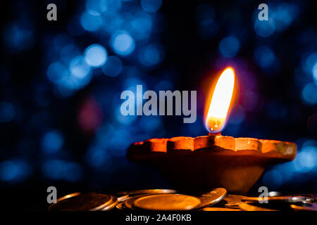 Low angle, closeup of a beautiful earthen lamp glowing in the dark with blue bokeh in the background, on a pile of coins in Diwali - prosperity concep Stock Photo
