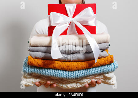 A woman holds a stack of warm knitted sweaters and a box with a gift on a gray wall background. Stock Photo
