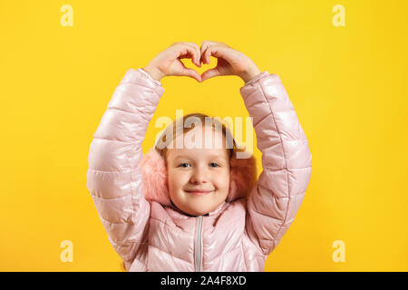 Cheerful little girl shows a heart with her fingers over her head. A child in a jacket and warm ear muffs on a yellow background. The concept of winte Stock Photo