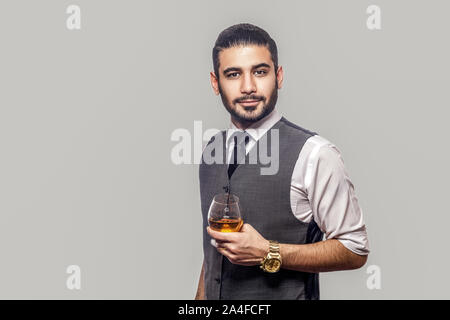 Portrait of handsome bearded brunette man in white shirt and waistcoat standing, holding glass of whiskey and looking at camera with serious face. ind Stock Photo