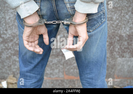 Arrested drug dealer in handcuffs with small heroin drug package on dark wall background. Illegal drug trafficking concept Stock Photo