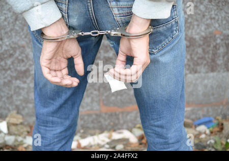 Arrested drug dealer in handcuffs with small heroin drug package on dark wall background. Illegal drug trafficking concept Stock Photo