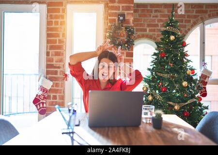 Beautiful woman sitting at the table working with laptop at home around christmas tree Posing funny and crazy with fingers on head as bunny ears, smil Stock Photo