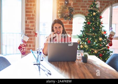 Beautiful woman sitting at the table working with laptop at home around christmas tree Pointing with hand finger to face and nose, smiling cheerful. B Stock Photo