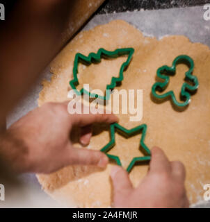male hands cut out gingerbread cookie in the form of a snowflake, Christmas tree, a man from raw dough on parchment baking paper on a dark background Stock Photo