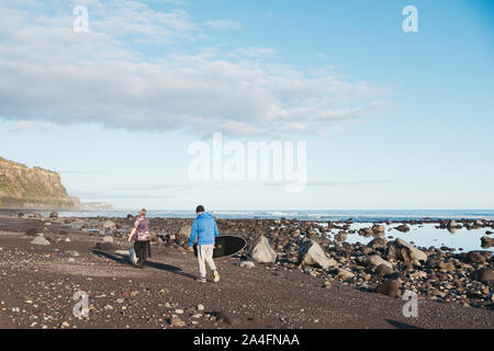 Father and son walking a dog down rocky beach carrying a surfboard Stock Photo