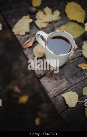 On a old wooden table in the autumn park is a cup with tea and coffee, scattered yellow leaves and pine cones. Top view, in blur. Autumn warm dark Stock Photo