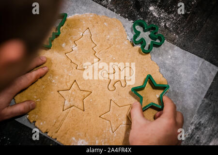 male hands cut out gingerbread cookie in the form of a snowflake, Christmas tree, a man from raw dough on parchment baking paper on a dark background Stock Photo