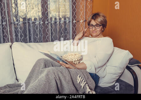 Middle aged woman reading book and eating popcorn while sitting on the couch at home Stock Photo